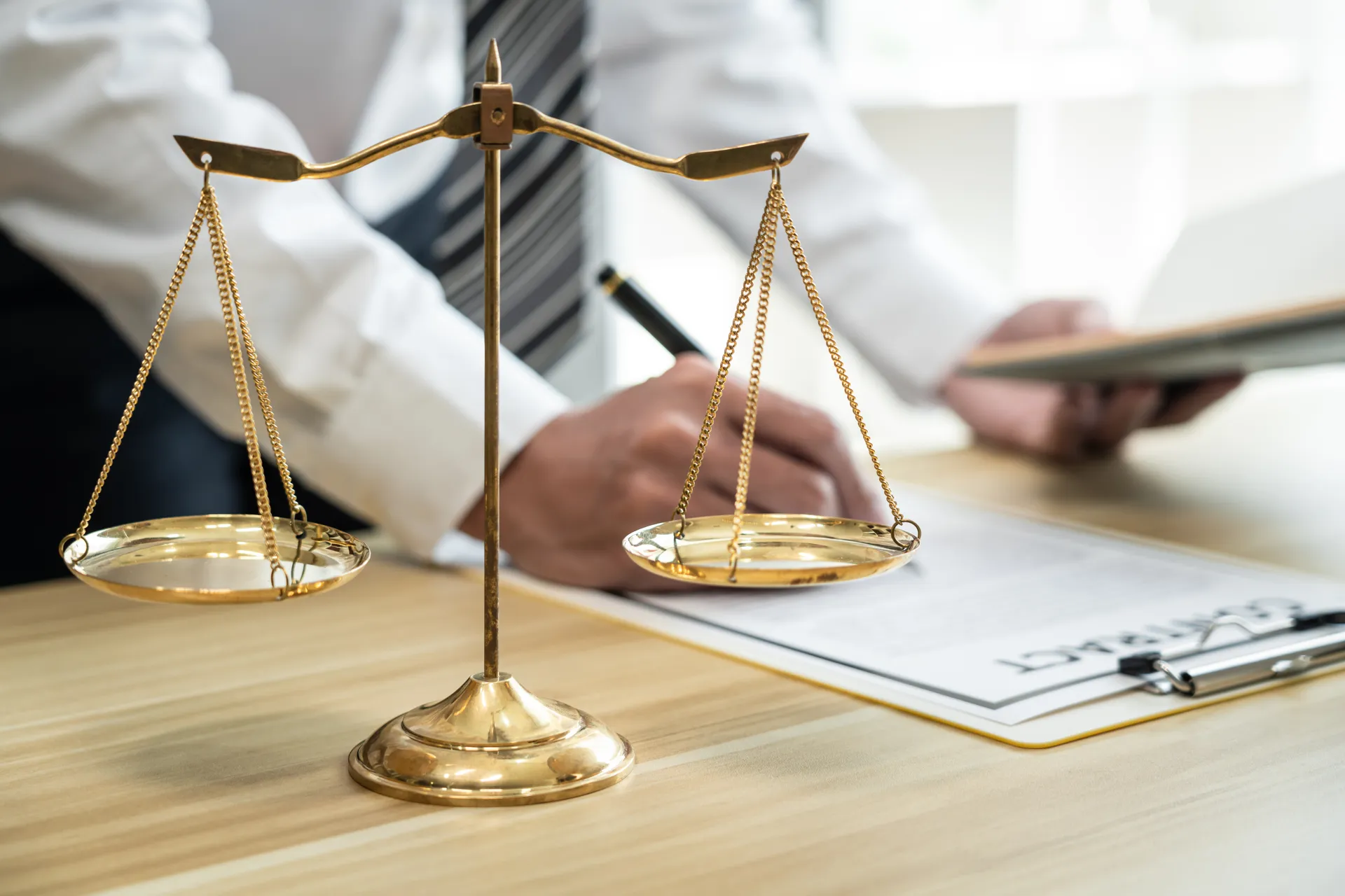 Balanza de la justicia en un escritorio, mientras un abogado firma documentos legales. La imagen simboliza el equilibrio y la equidad en el ámbito jurídico.