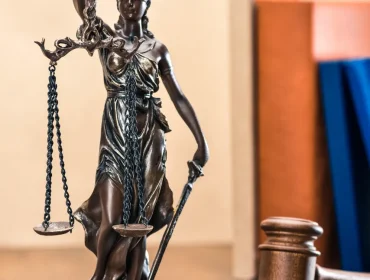 Estatua de la Justicia, representada por una figura femenina con venda en los ojos, sosteniendo una balanza y una espada, en un entorno de oficina legal con un martillo de juez al fondo.