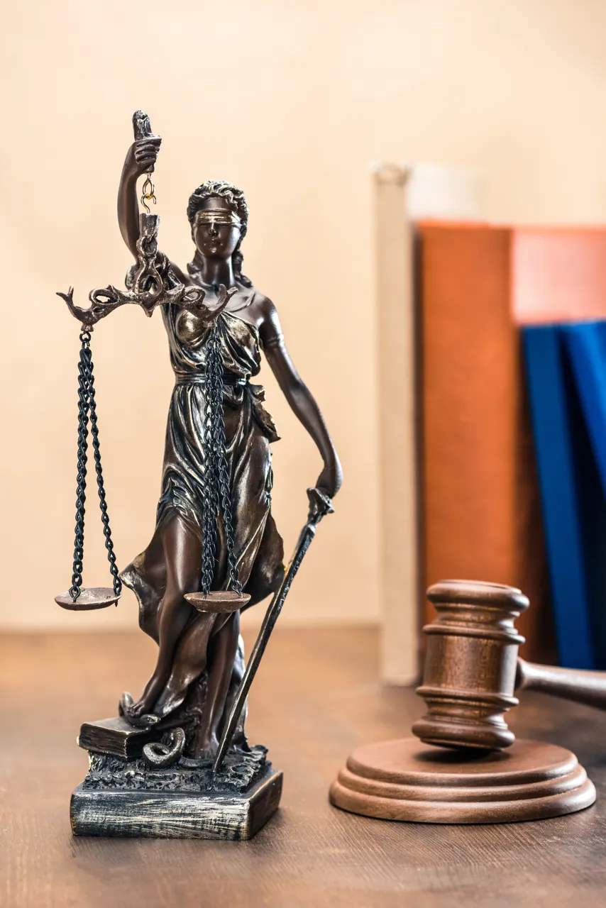 Estatua de la Justicia, representada por una figura femenina con venda en los ojos, sosteniendo una balanza y una espada, en un entorno de oficina legal con un martillo de juez al fondo.