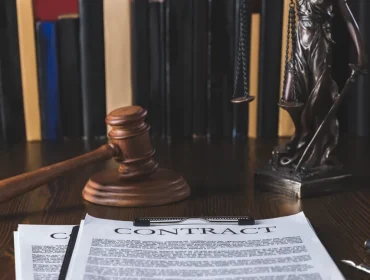 Imagen que muestra documentos de contrato sobre una mesa de madera, junto a un martillo de juez y una figura de la justicia, con una estantería de libros legales al fondo. Ideal para temas legales y jurídicos.